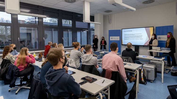 Blick von hinten in einem modernen Seminarraum, vorne stehen drei Studierende vor einem großen Bildschirm und halten eine Präsentation.