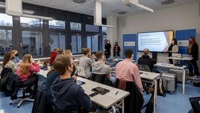 Blick von hinten in einem modernen Seminarraum, vorne stehen drei Studierende vor einem großen Bildschirm und halten eine Präsentation.