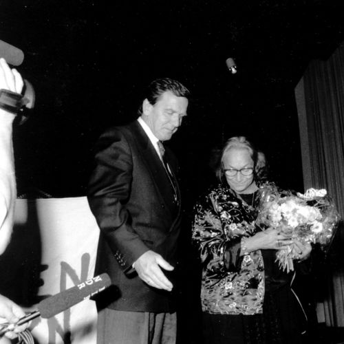 Das Bild zeigt den damaligen niedersächsischen Ministerpräsidenten Gerhard Schröder mit Rosalinde von Ossietzky-Palm. Sie stehen auf einer Bühne, sie hält einen Blumenstrauß in der Hand. Schröder weist ihr den Weg von der Bühne. Kameras beobachten die Szene.