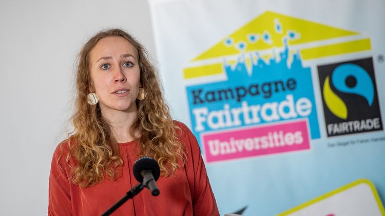 Young woman standing in front of a microphone, campaign poster in the background.