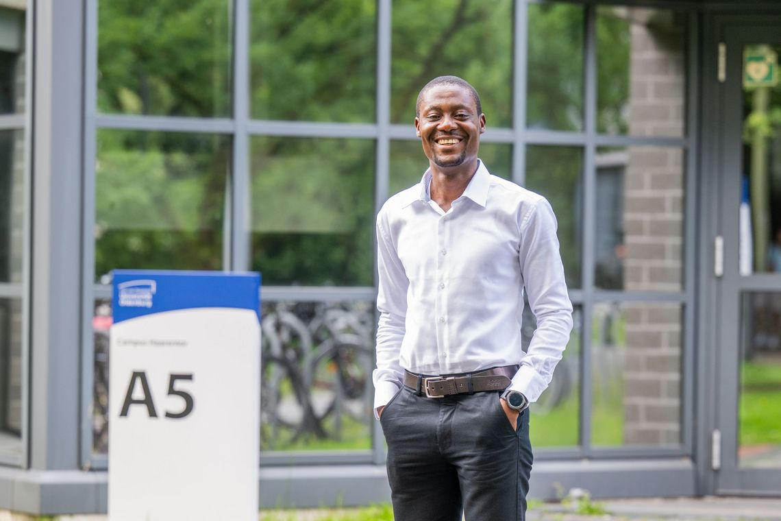 Emmanuel Asane-Otoo steht vor dem Eingang von Gebäude A 5. Er lächelt in die Kamera und hält die Hände in den Hosentaschen. Im Hintergrund spiegeln sich Fahrräder in den Fensterscheiben.