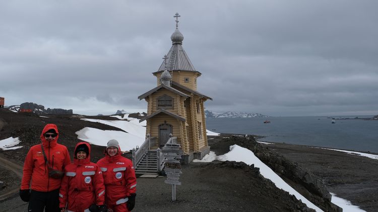 Gruppenbild Jens Meyerjürgens, Isabell Schlangen und Lina Holthusen. Das Team trägt rote Polaranzüge, im Hintergrund ist die kleine orthodoe Kirche auf King George Island sowie eine Meeresbucht zu sehen. 