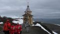 Group photo of Jens Meyerjürgens, Isabell Schlangen and Lina Holthusen. The team is wearing red polar suits; the small Orthodox church on King George Island and a bay can be seen in the background. 