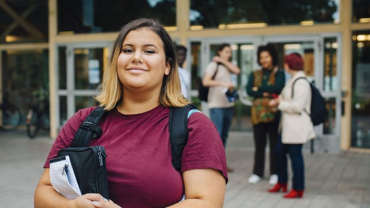 Studentin vor Gebäude auf dem Universitätscampus guckt in die Kamera,hinter ihr unterhalten sich weitere Studierende