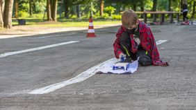 Ein Junge hockt auf dem Schulhof, tunkt eine Rolle in eine Farbpalette, um eine gerade, weiße Linie auf das Pflaster zu malen.