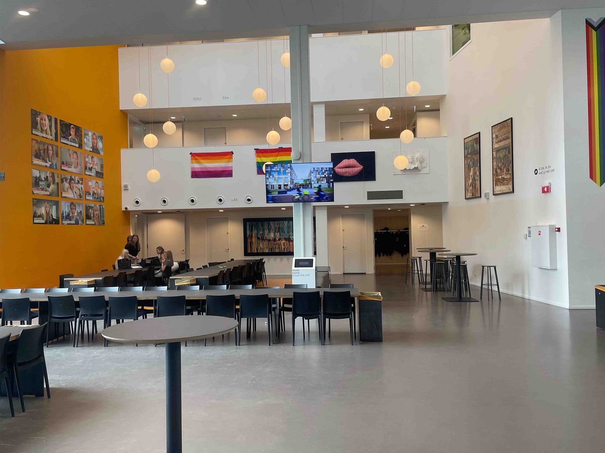 Teaching Across Borders Ein großer, heller Flur in der Schule Stad&Esch mit Porträts, Kunstwerken und Regenbogenflaggen. Im Mittelpunkt ein großer Bildschirm mit Video.