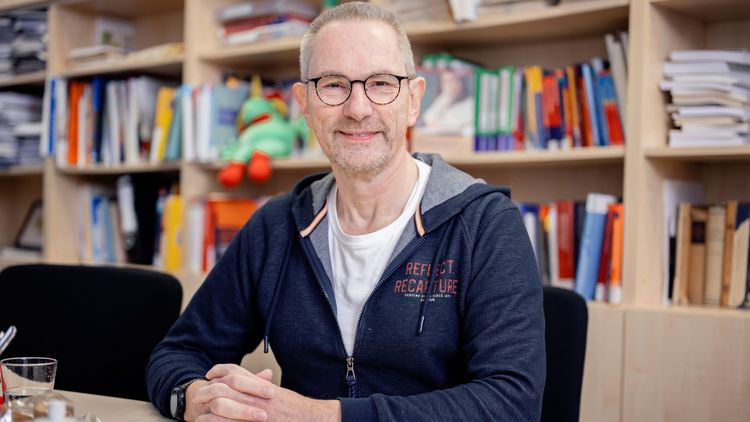 Das Bild zeigt Clemens Hillenbrand. Er sitzt an einem Tisch vor einem Bücherregal und schaut in die Kamera.