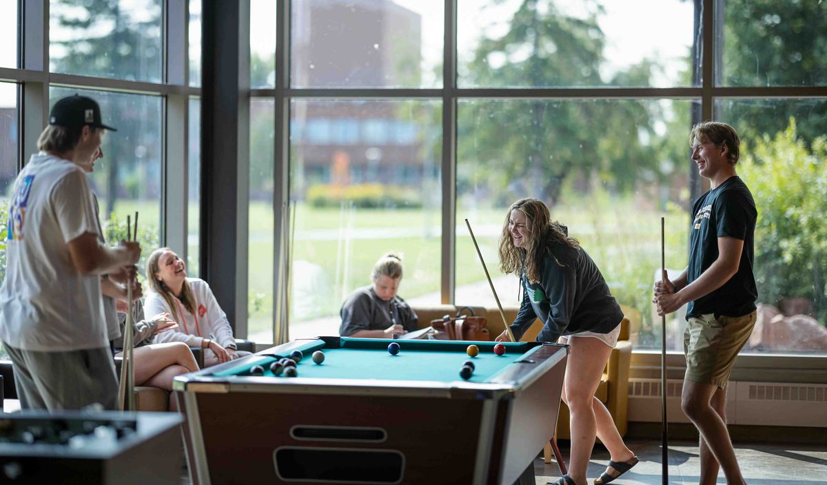 Studierendenleben Studierende spielen Billard in einem hellen Aufenthaltsraum mit großen Fenstern.