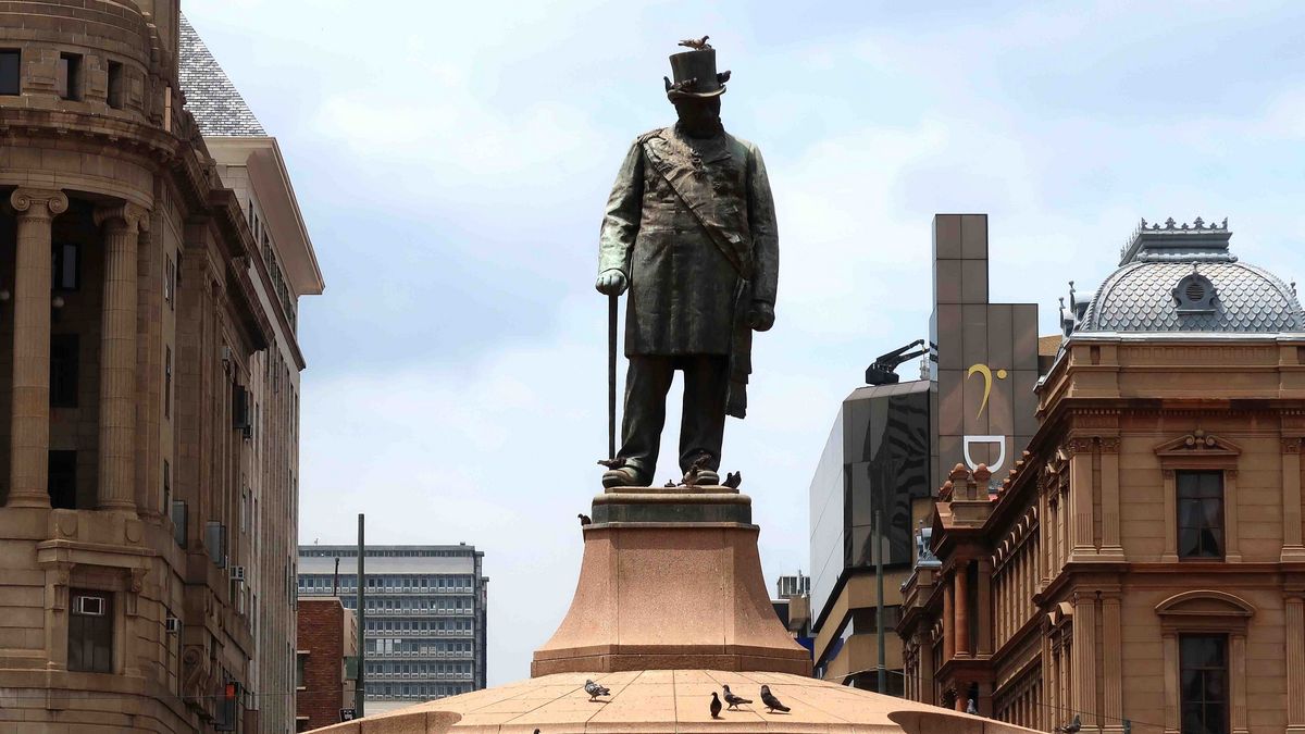 Das Bild zeigt eine Bronzestatue von Paul Kruger, einem Präsidenten der Südafrikanischen Republik (Transvaal). Die Statue zeigt Kruger in historischer Kleidung, darunter einen Zylinder und einen langen Mantel, und er hält einen Spazierstock. Die Statue steht auf einem hohen Sockel inmitten einer städtischen Umgebung mit historischen und modernen Gebäuden. Im Hintergrund sind weitere Gebäude und ein leicht bewölkter Himmel zu sehen. Einige Vögel sind auf dem Sockel und in der Nähe der Statue zu erkennen.