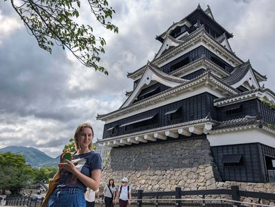 Burg Kumamoto Burg Kumamoto, eine mehrstöckige Burg mit dunklen Holzaußenwänden und einer Person im Vordergrund.