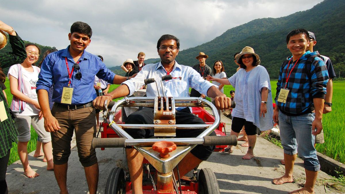 Das Bild zeigt eine Gruppe von (internationalen) Studierenden und möglicherweise Dozierenden der National Dong Hwa University (NDHU) bei einer Exkursion in eine ländliche Gegend. Im Vordergrund sitzt ein Mann auf einem kleinen, roten Traktor, während andere Personen um ihn herum stehen und lächeln. Die Gruppe befindet sich in der Nähe von Reisfeldern und einer grünen Landschaft mit Bergen im Hintergrund. Es scheint sich um eine kulturelle oder erlebnisorientierte Aktivität der Universität zu handeln.