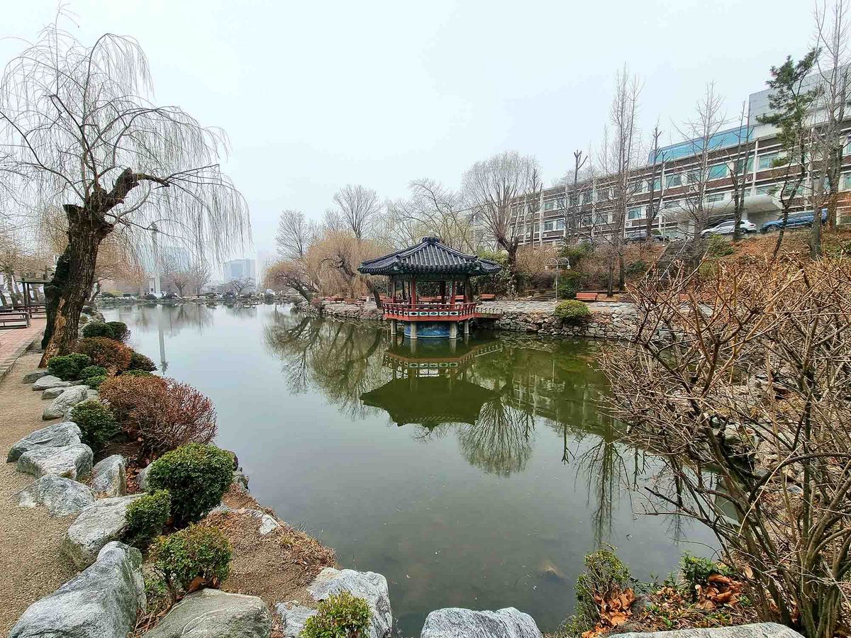 Das Bild zeigt einen kleinen Teich oder See auf dem Campus der Inha University. Ein traditioneller, überdachter Pavillon steht am Ufer des Sees, der sich im Wasser spiegelt. Bäume säumen das Ufer, und im Hintergrund ist ein Gebäude zu sehen.