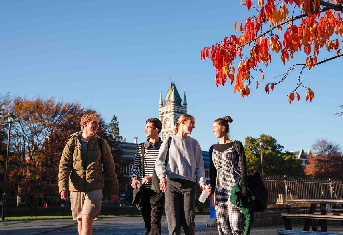 University of Otago Vier Studierende gehen lächelnd über einen Campusweg. Im Hintergrund der Glockenturm der University of Otago.
