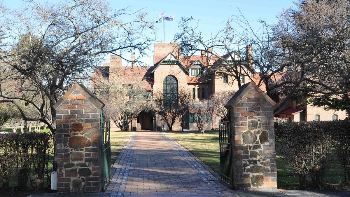 Das Bild zeigt einen historischen Gebäudekomplex auf dem Campus der University of New England. Das Gebäude ist aus rotem Backstein gebaut und weist eine beeindruckende Architektur mit Türmen und Bögen auf. Vor dem Gebäude befindet sich ein gepflasterter Weg, der von steinernen Pfeilern mit einem grünen Tor flankiert wird. Im Hintergrund sind Flaggen zu sehen und Bäume umrahmen die Szene.