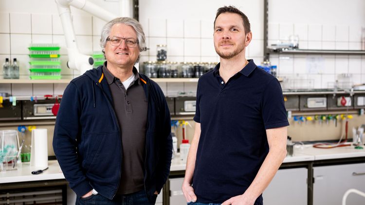 Zwei Männer stehen in einem Labor vor einem Arbeitsbereich mit Schränken, einer Arbeitsplatte und verschiedenen Geräten. 