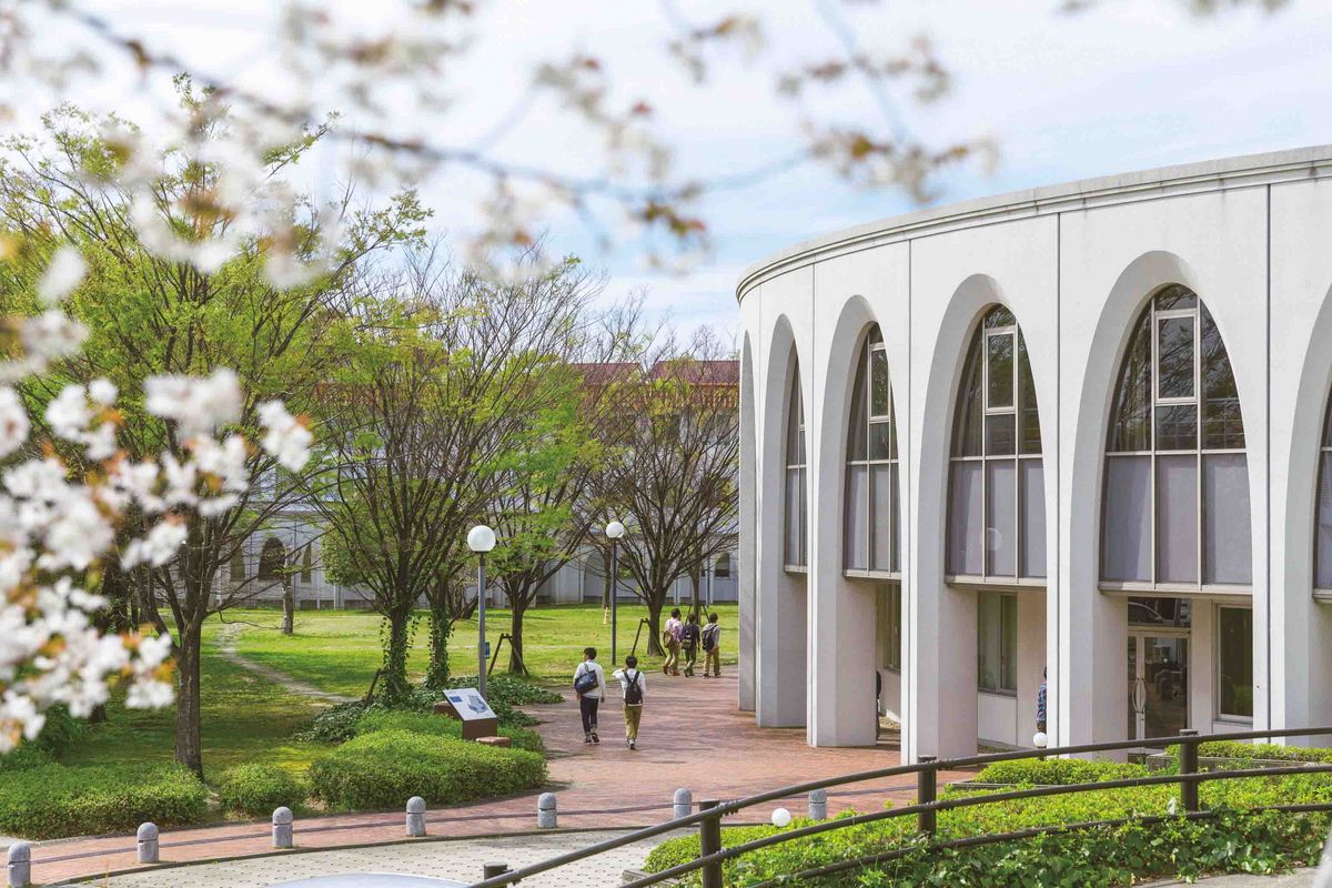 Ein Gebäude auf dem Nagoya Campus der Chukyo Universität mit einer gebogenen Fassade und Arkaden. Im Vordergrund befindet sich ein gepflegter Rasen mit Bäumen und Wegen. Einige Personen gehen auf dem Weg entlang. Blütenzweige sind im Vordergrund zu sehen.