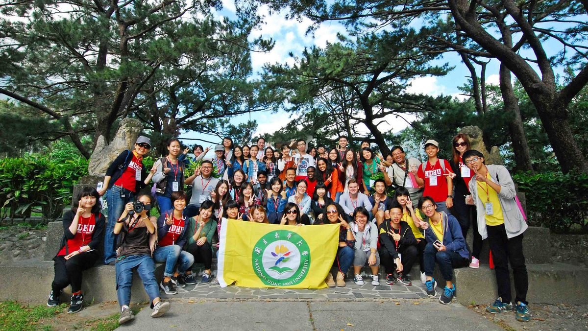 Das Bild zeigt eine große Gruppe von Studierenden und auch Mitarbeitenden der National Dong Hwa University (NDHU) bei einer Wanderung oder einem Ausflug in einer bewaldeten Umgebung. Die Gruppe posiert für ein Gruppenfoto vor einem Stoffbanner mit dem Logo der NDHU. Die Szene vermittelt eine gemeinschaftliche und freundliche Atmosphäre.