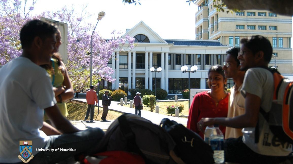  Mehrere Studierende sitzen auf einer Bank auf dem Campus der University of Mauritius und unterhalten sich. Im Hintergrund befindet sich ein Gebäude mit weißer Fassade und Säulen, umgeben von Bäumen mit lilafarbenen Blüten. Einige weitere Personen gehen auf dem Campus entlang.