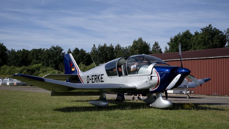 Ein kleines Flugzeug steht auf einer Wiese vor einem Hangar, im HIntergrund Bäume und blauer Himmel..