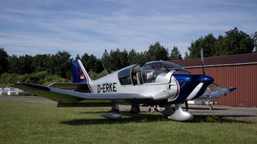 Ein kleines Flugzeug steht auf einer Wiese vor einem Hangar, im HIntergrund Bäume und blauer Himmel..
