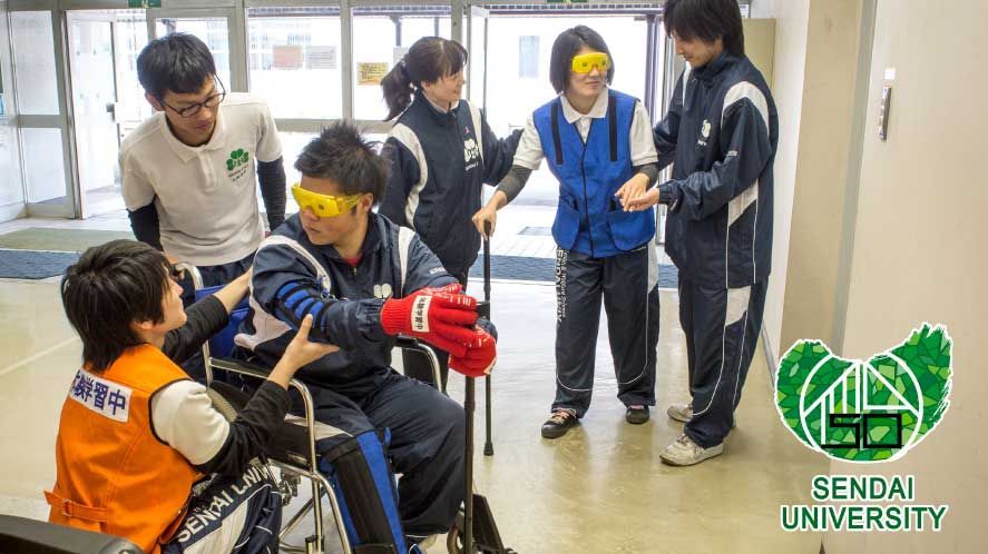 Das Bild zeigt eine Gruppe von Studierenden der Sendai University, die an einer Übung teilnehmen, bei der eine Person im Rollstuhl geführt wird. Mehrere Studierende in farbigen Westen und Jacken stehen um eine Person im Rollstuhl und bieten Unterstützung und Anleitung. Die Brillen, die einige der Studierenden tragen, sind sogenannte "Simulationsbrillen". Sie sind dazu gedacht, die Sehbehinderung zu simulieren, die Menschen mit bestimmten Augenerkrankungen erleben. Die Szene spielt in einem hellen Innenraum, möglicherweise einem Flur oder einer Halle der Universität und vermittelt einen Eindruck von Inklusion und Unterstützung.