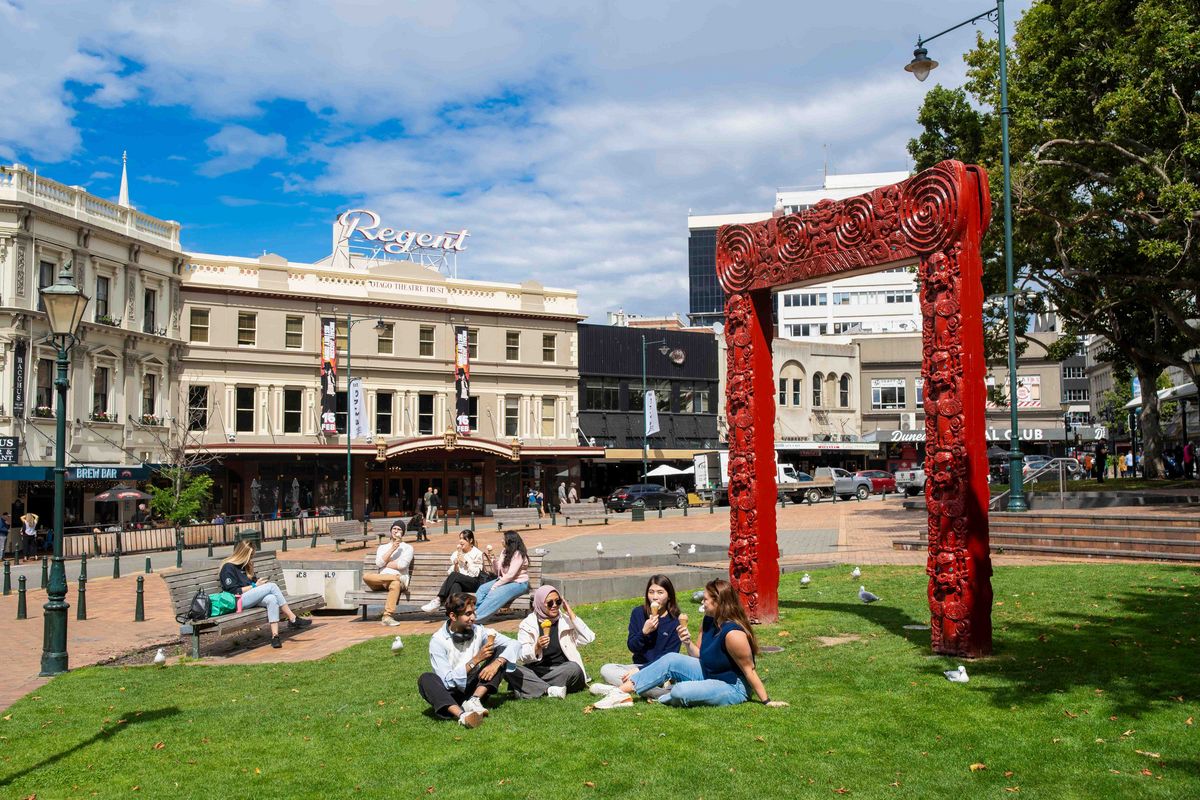 Das Bild zeigt eine belebte Szene im Herzen von Dunedin, Neuseeland, mit dem historischen Regent Theatre und einem beeindruckenden Māori-Schnitzwerk im Vordergrund. Studierende und Einheimische entspannen sich auf dem grünen Rasen vor dem Theater, genießen die Sonne und unterhalten sich. Die Szene vermittelt ein Gefühl von Gemeinschaft und die lebendige Atmosphäre.