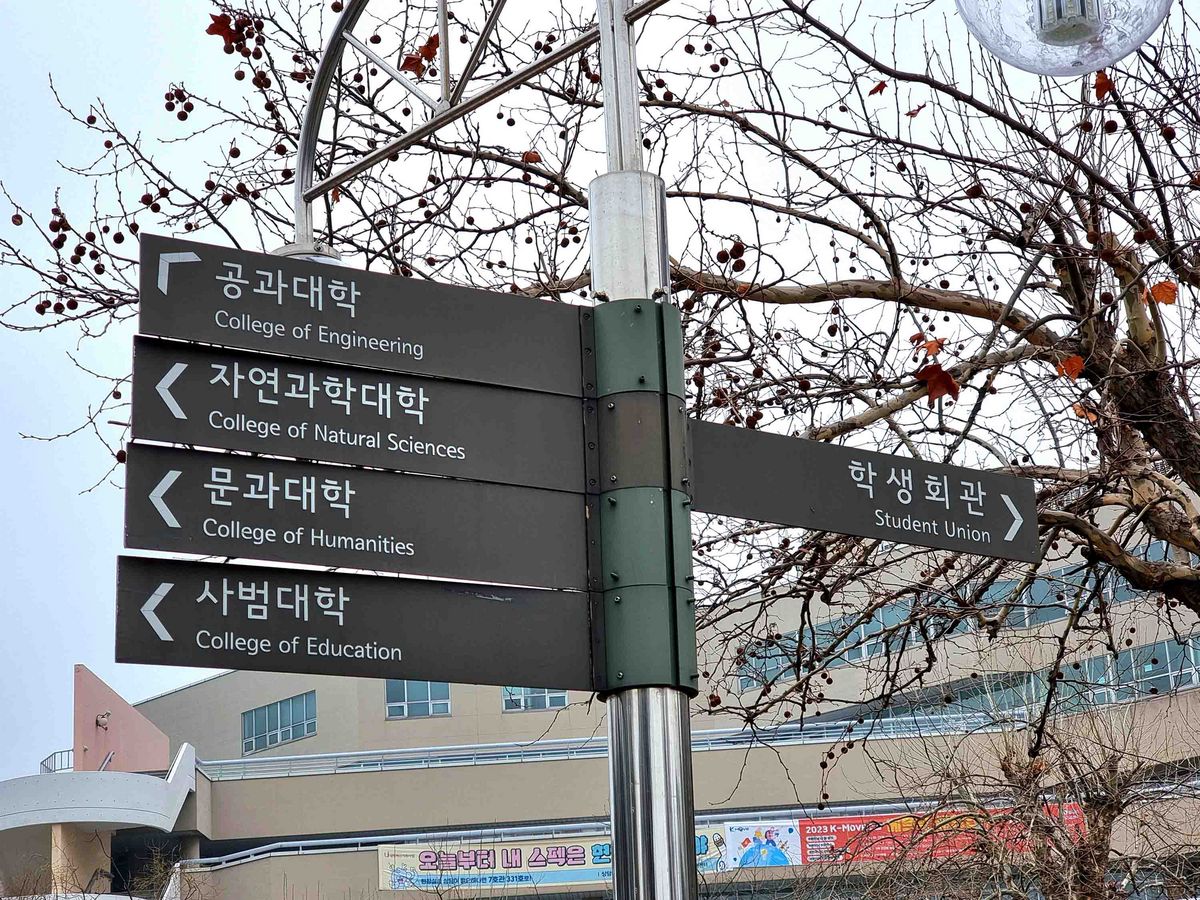 Das Bild zeigt ein Schild mit Wegweisern auf dem Campus der Inha University. Das Schild weist in verschiedene Richtungen zu den Colleges für Ingenieurwesen, Naturwissenschaften, Geisteswissenschaften und Pädagogik sowie zur Studentenunion. Im Hintergrund sind Gebäude und kahle Bäume zu sehen.
