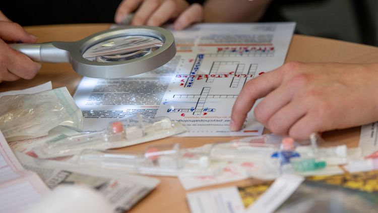 Nahaufnahme einer Lupe, die über ein Kreuzworträtsel gehalten wird. Darum herumg sind medizinische Materialien zu sehen, etwa Spritzen und ein Rezept.