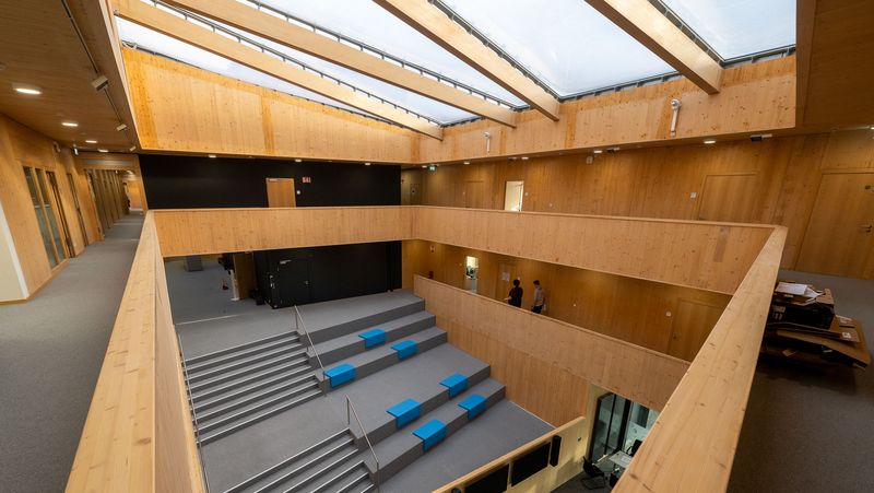 Blick von der oberen Galerie auf den Inneraum des Gebäudes. Durch das Dach kommt Licht herein, im Inneren ist alles mit Holz verkleidet und neben der Treppe gibt es Absätze, wo man sich hinsetzen kann. 