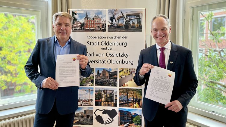 Zwei Männer stehen vor einem Aufsteller und halten ein Blatt Papier in die Kamera. 