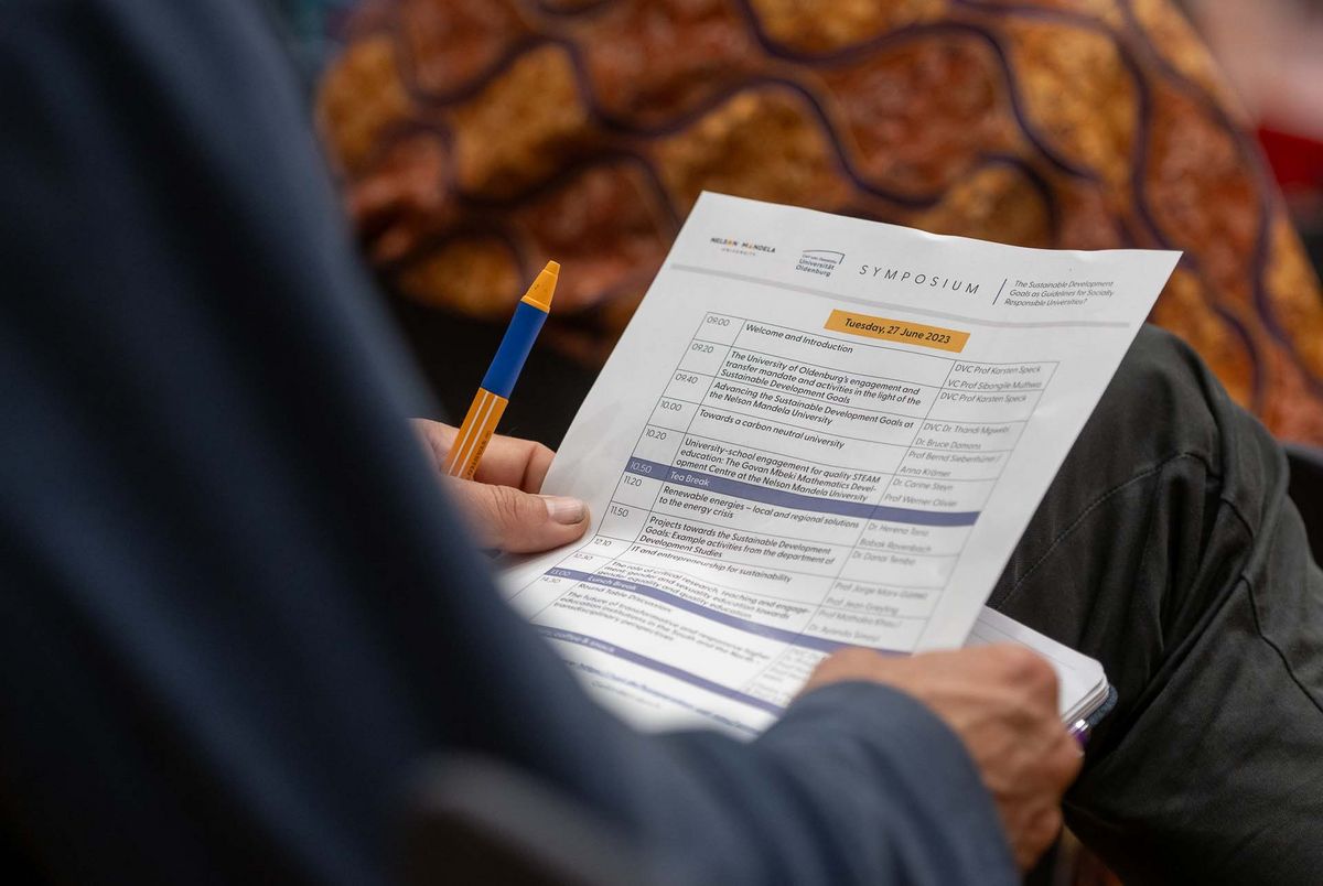 Symposium Das Bild zeigt einen Programmplan für ein Symposium, das am 27. Juni 2023 stattfindet. Der Plan wird von den Händen einer Person gehalten und zeigt verschiedene Vorträge und Präsentationen zu Themen wie nachhaltiger Entwicklung, Bildung und den Zielen der Nelson Mandela University.