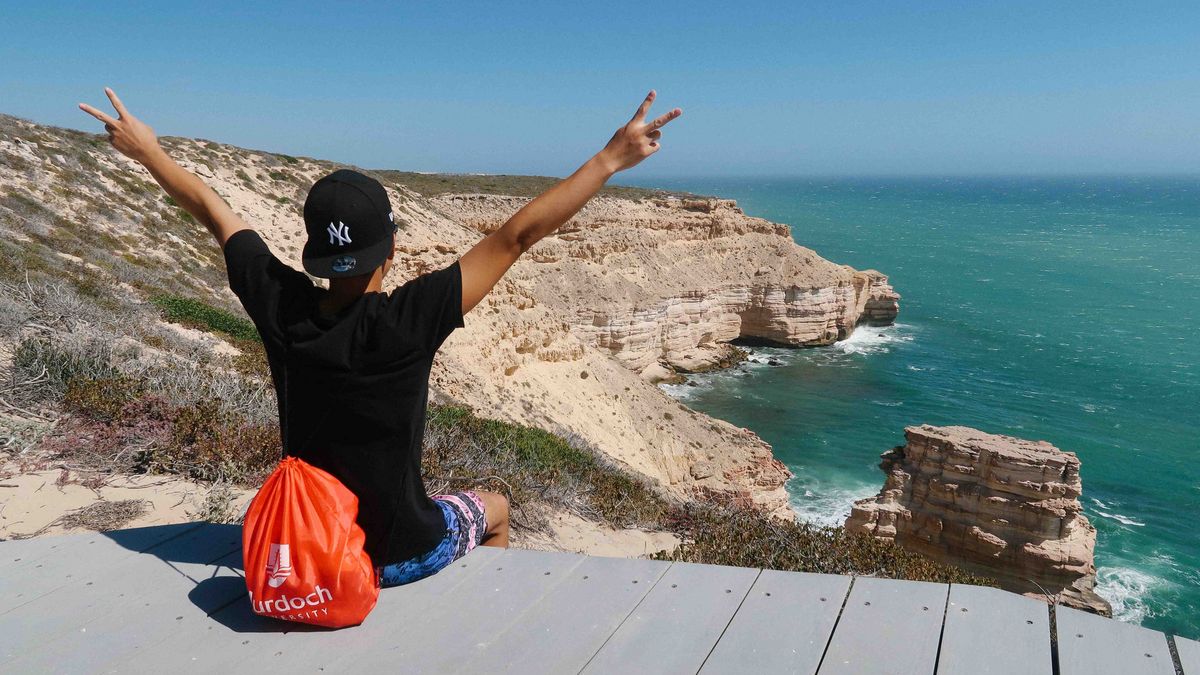 Das Bild zeigt eine Person, die mit ausgebreiteten Armen auf einer Holzplattform am Rande einer Klippe steht. Die Person trägt eine Kappe und ein T-Shirt und blickt auf das Meer. Im Hintergrund erstreckt sich eine zerklüftete Küstenlinie mit weißen Klippen und türkisfarbenem Wasser. Eine Murdoch University Tragetasche liegt direkt hinter der Person. Die Szene vermittelt ein Gefühl von Freiheit, Freude und Abenteuer.