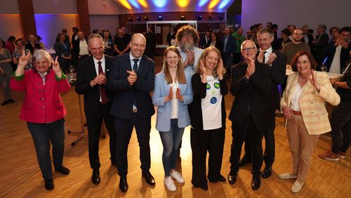 Das Bild zeigt die jubelnden Mitglieder des Präsidiums, die Cluster-Sprecher und weitere Personen, die in Feierlaune sind. Einige heben die Arme, klatschen oder machen das V-Zeichen.