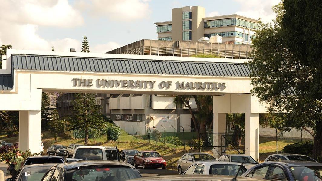 Der Torbogen mit der Aufschrift "The University of Mauritius" ist im Vordergrund zu sehen, während sich Autos auf der Zufahrtsstraße zum Campus befinden. Im Hintergrund erstreckt sich ein Gebäude der Universität.