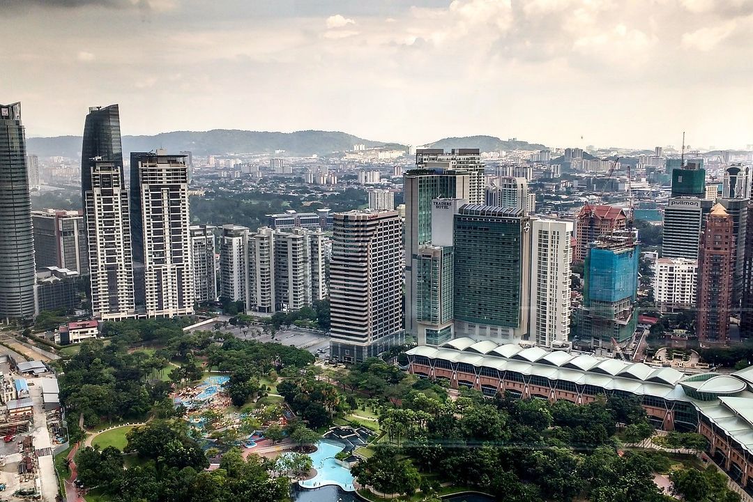 Das Bild zeigt eine Panoramaaufnahme der Stadt Kuala Lumpur, Malaysia, mit ihren hohen Wolkenkratzern. Im Vordergrund befindet sich ein Park mit Bäumen und einem Schwimmbad. Im Hintergrund erstreckt sich die Stadt bis zu den Hügeln am Horizont. Der Himmel ist bewölkt. Die Szene vermittelt einen Eindruck von urbanem Leben und moderner Architektur.