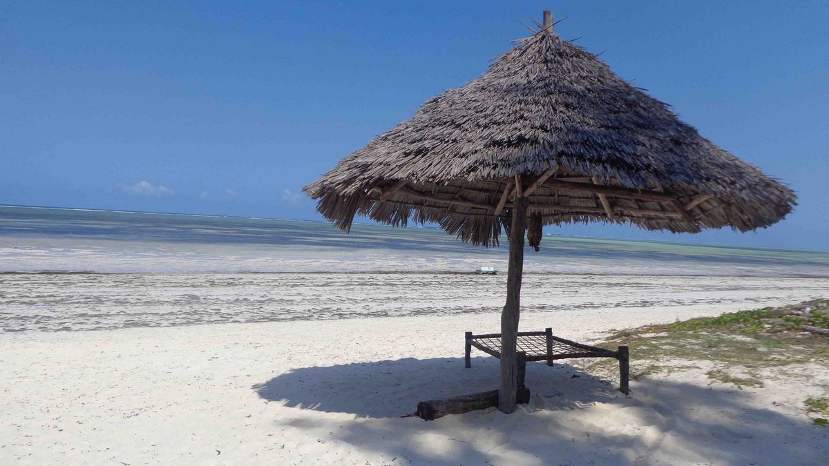 Strand in Dar es Salaam in traditioneller Strohhut-Pavillon am Strand von Dar es Salaam, Tansania. Im Hintergrund das türkisfarbene Meer unter einem blauen Himmel.