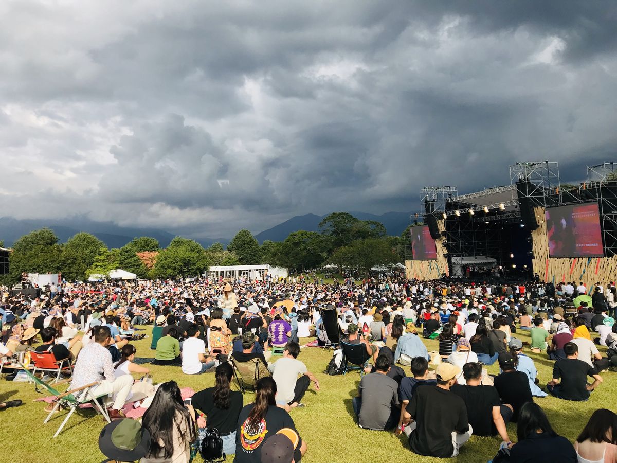 Das Bild zeigt eine riesige Menschenmenge bei einem Konzert oder Festival auf einer grünen Wiese unter einem dramatischen, bewölkten Himmel.