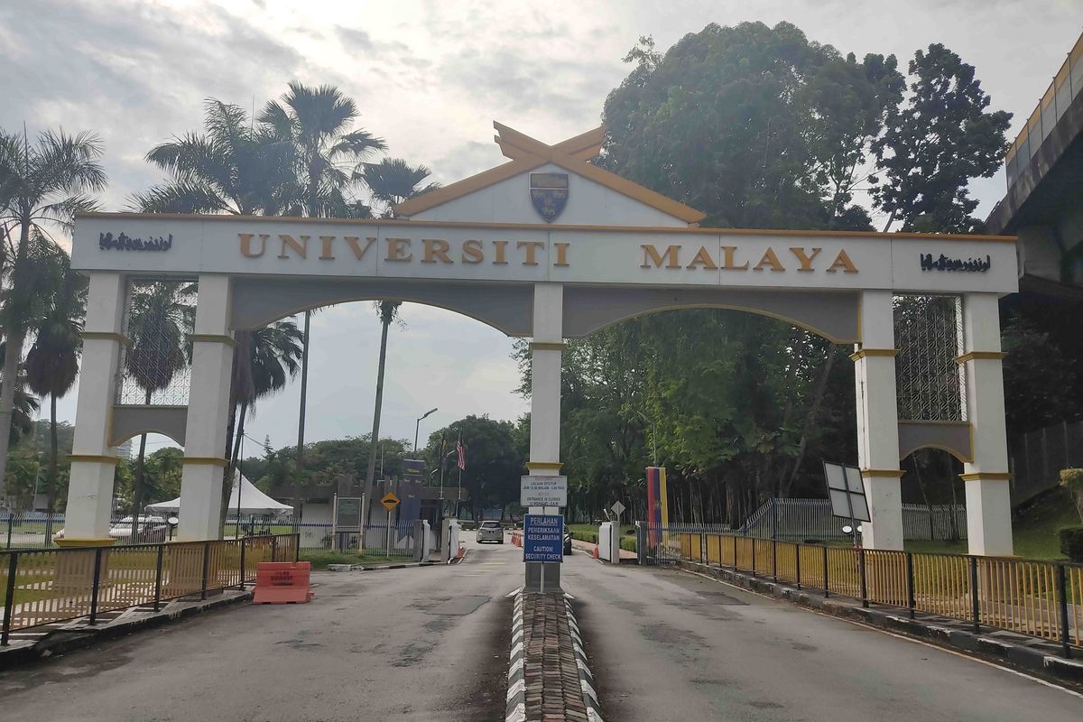 Das Bild zeigt das Haupteingangstor der University of Malaya in Kuala Lumpur, Malaysia. Über dem Torbogen befindet sich der Schriftzug "Universiti Malaya" in gelber Schrift auf weißem Hintergrund. Das Tor ist von grüner Vegetation und Bäumen umgeben. Im Hintergrund sind weitere Gebäude und ein bewachter Kontrollpunkt zu sehen. Die Szene vermittelt einen Eindruck von akademischer Tradition und Sicherheit.