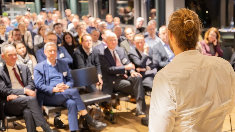Ein Mann im weißen Hemd und zusammengebundenen Haaren ist von hinten zu sehen. Er hält einen Vortrag vor einem größeren Publikum. 