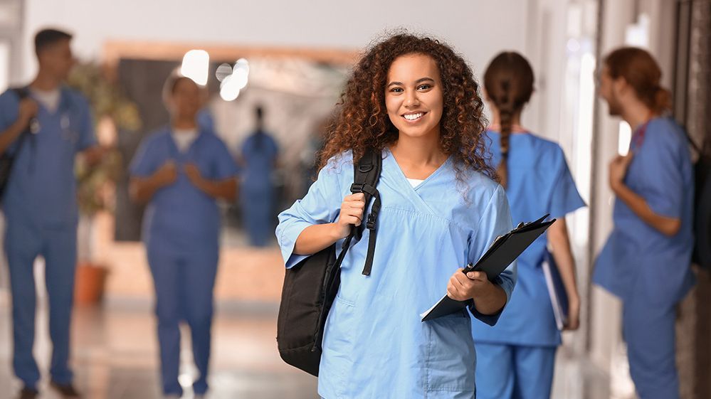 Gesundheit und Soziales Eine junge Frau in Arbeitskleidung befindet sich in einer medizinischen Einrichtung. Im Hintergrund sind weitere Kolleginnen und Kollegen zu erkennen.