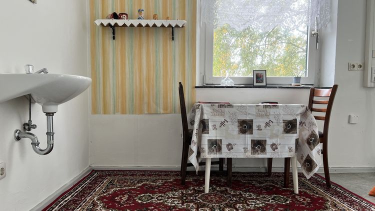 Das Foto gibt Einblick in ein inszeniertes Esszimmer, das optisch an sterotypische Esszimmer russlanddeutscher-Familien angelehnt ist. Es liegt ein roter, gemusterter Teppich auf dem Boden. Darauf steht ein Tisch mit Matroschkapuppen. Alles wirkt altmodisch.