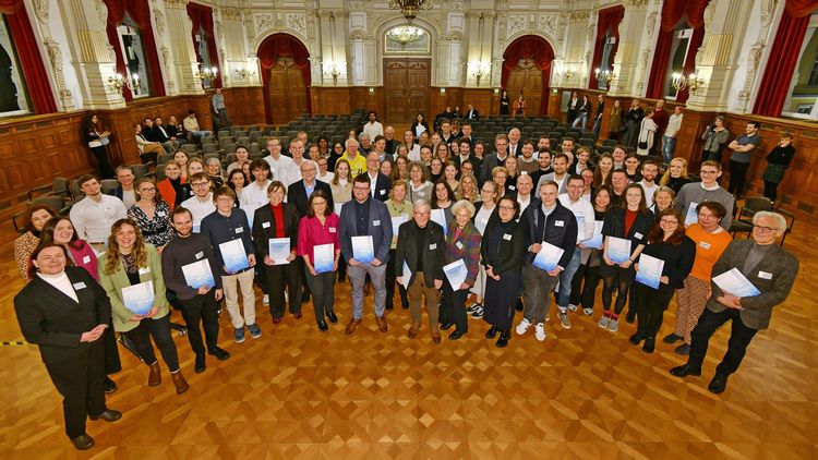 Das Bild zeigt die Geförderten sowie ihre Förderinnen und Förderer auf einem großen Gruppenbild. Sie schauen in die Kamera und zeigen ihre Urkunden. Die Aufnahme wurde im prächtigen Festsaal des Oldenburger Schlosses gemacht. In ihm befinden sich stuckverzierte Wände und Decken, Kronleuchter und rote Vorhänge.