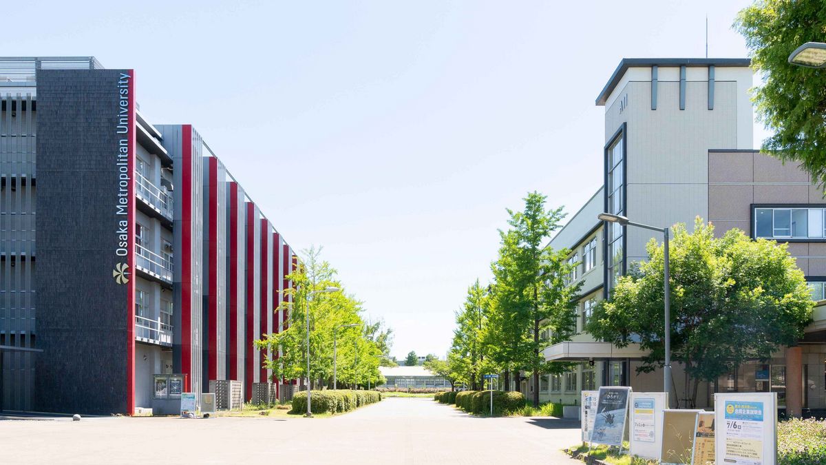 Das Bild zeigt einen modernen Gebäudekomplex der Osaka Metropolitan University, der aus grauen und roten Segmenten besteht. Der graue Teil des Gebäudes ist hoch und schmal, mit vertikalen Linien und dem Schriftzug "Osaka Metropolitan University". Vor dem Gebäude verläuft ein asphaltierter Weg, gesäumt von Bäumen und Plakatständern mit Ankündigungen und Informationen. Die Szene wirkt friedlich und einladend, mit viel Tageslicht und Grünflächen
