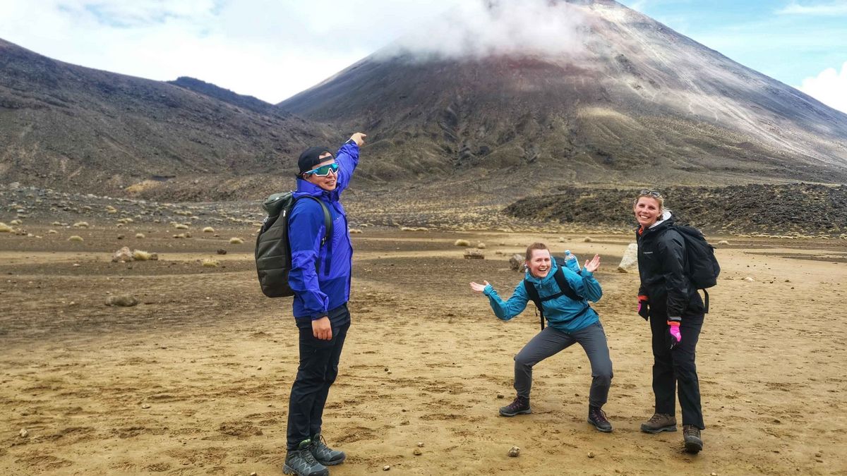 Das Bild zeigt drei lächelnde Personen auf einem kargen, vulkanischen Gelände. Im Hintergrund erhebt sich ein großer, kegelförmiger Vulkan, dessen Spitze von Wolken verdeckt ist. Die Personen tragen Wanderkleidung und Rucksäcke und scheinen einen Ausflug zu genießen.