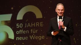 Ralph Bruder mit Mikrofon auf der Bühne, hinter ihm das Jubiläumslogo "50 Jahre offen für neue Wege". 