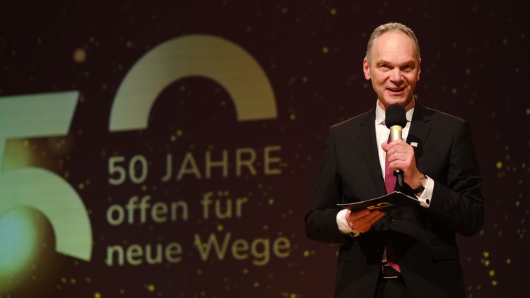 Ralph Bruder mit Mikrofon auf der Bühne, hinter ihm das Jubiläumslogo "50 Jahre offen für neue Wege". 
