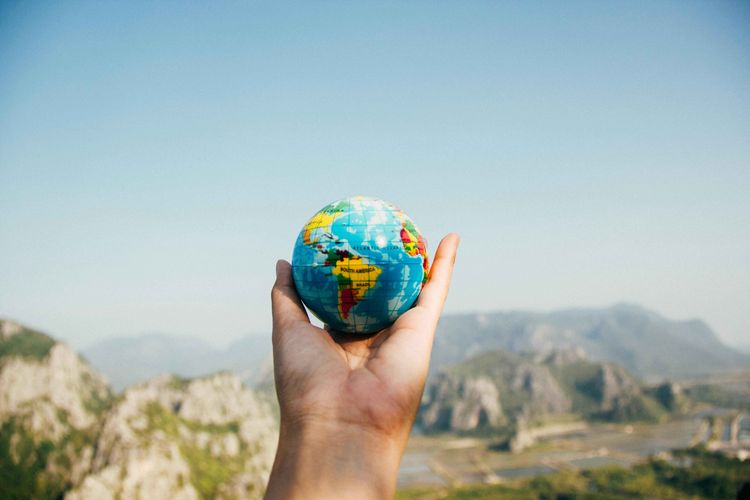 Erdkugel Eine Hand hält eine kleine Weltkugel vor einer verschwommenen Landschaft mit Bergen. Die Weltkugel zeigt Kontinente und Ozeane in blauen und grünen Farbtönen. Die Aufnahme vermittelt ein Gefühl von Globalität, Reisen und der Verbindung zwischen Mensch und Welt.