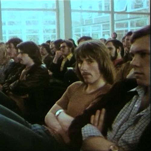 Das Bild ist ein Screenshot aus einem Imagefilm der Uni. Es zeigt Studierende, die in der Aula in A 11 sitzen und mit verschränkten Armen einem Vortrag lauschen. Einer der Männer schaut leicht genervt in die Kamera.