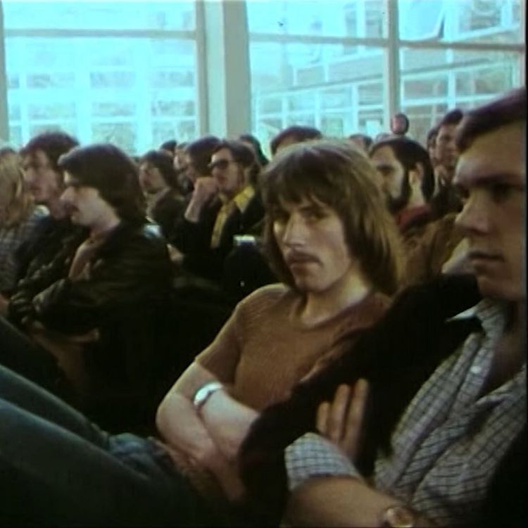 Das Bild ist ein Screenshot aus einem Imagefilm der Uni. Es zeigt Studierende, die in der Aula in A 11 sitzen und mit verschränkten Armen einem Vortrag lauschen. Einer der Männer schaut leicht genervt in die Kamera.