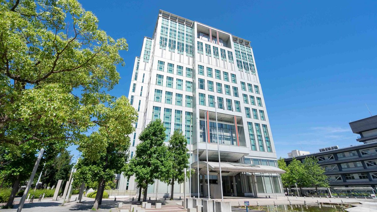 Das Bild zeigt einen hohen, modernen Wolkenkratzer der Osaka Metropolitan University, der sich gegen einen klaren blauen Himmel erhebt. Das Gebäude hat eine helle Fassade mit vielen rechteckigen Fenstern und ist durch horizontale Linien gegliedert. Vor dem Gebäude befindet sich ein kleiner Platz mit Bäumen, Bänken und einem kleinen Wasserbecken, der einen Kontrast zur modernen Architektur bildet.